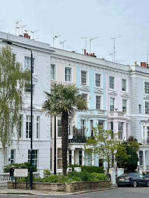 The image depicts a row of white Victorian-style residential buildings with detailed architectural features, including decorative moldings and bay windows, situated in Notting Hill. In front of the buildings, there is a small garden area with neatly maintained shrubs, a tall palm tree, and a black iron fence. A black parking meter and a car are visible parked along the street. The paved sidewalk runs parallel to the street, with a lamppost positioned near the garden. The scene is illuminated with natural daylight, highlighting the clean and well-maintained exterior surfaces of the buildings. This setting emphasizes the importance of careful property maintenance, akin to professional domestic cleaning and surface sanitisation provided by Notting Hill Carpet Cleaning, suited for precise surface cleaning and deep cleaning tasks in residential environments.