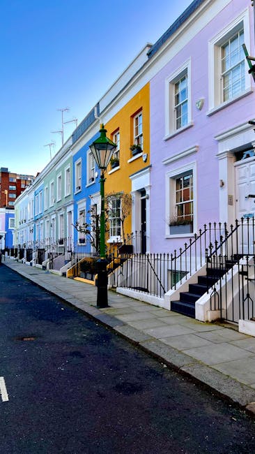 A row of brightly painted terraced houses on a residential street in Notting Hill, featuring pastel-colored facades in shades of blue, yellow, and lavender. Each house has a small front door with white trim, black wrought iron railings along the front steps, and potted plants near the entrances. A vintage-style green street lamp stands on the pavement, and the sidewalk is clean with neatly maintained stairs and pathways. The sky above is clear and blue, enhancing the vibrant and tidy appearance of the neighborhood. This exterior scene exemplifies the charming, well-kept character typical of Notting Hill and reflects a focus on maintaining pristine property exteriors, aligned with professional cleaning standards from Notting Hill Carpet Cleaning.