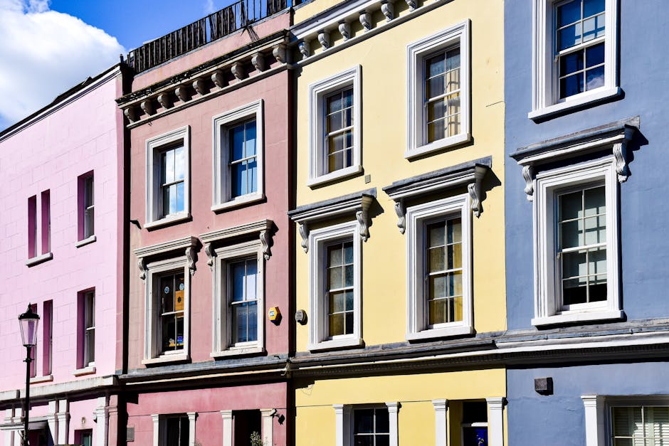 A row of three residential buildings painted in pastel colors—pink, yellow, and blue—located in Notting Hill. The buildings feature white-framed sash windows with decorative cornices and lintels, set against a partly cloudy sky. The pink building has three windows on the scaffolded upper floor and a streetlamp visible on the left. The yellow building displays a clean, well-maintained facade with neatly aligned windows and window sills. The blue building also shows similar white-framed windows and architectural details. The overall scene depicts an area that would benefit from professional surface cleaning and exterior maintenance, such as that provided by Notting Hill Carpet Cleaning, to enhance appearance and hygiene.