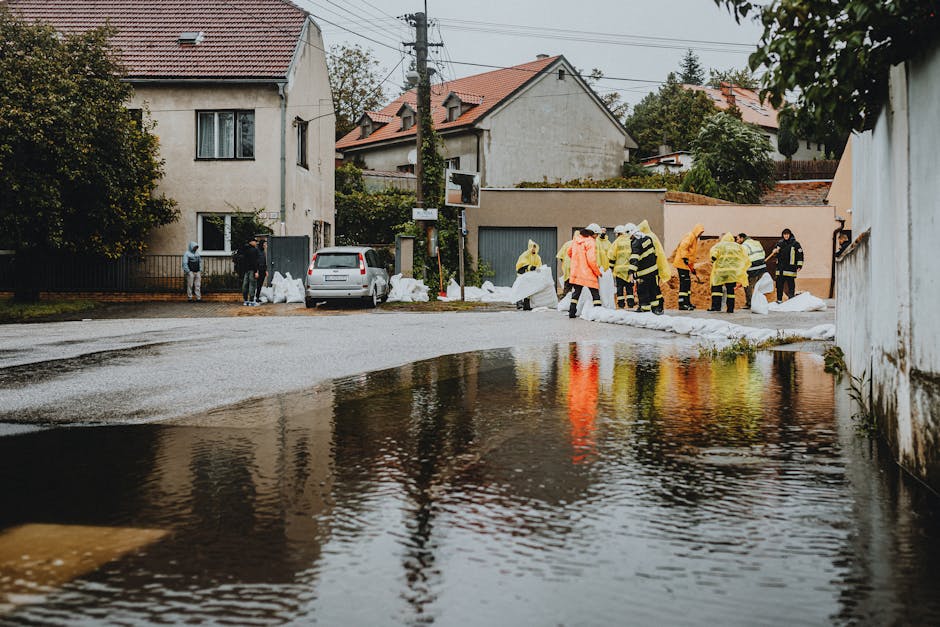 An outdoor scene during daylight showing a group of emergency response workers in high-visibility jackets and protective gear, standing on a residential street next to a white car and several sandbags stacked against a garage. The street is partially flooded with water reflecting trees and houses, which feature various materials including wood, brick, and stucco. The background includes detached houses with pitched roofs, several windows, and surrounding greenery. The scene is illuminated by natural daylight, and the wet pavement indicates recent flooding. Notting Hill Carpet Cleaning technicians recommend thorough surface cleaning and sanitisation following flood events to restore hygiene and safety in residential properties.
