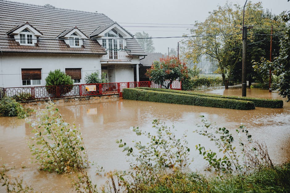 A flooding scene in a residential area of Pembridge Villas, Notting Hill, showing a house with a white exterior, small dormer windows, and a grey tiled roof. The front garden is submerged in muddy water, with a trimmed hedge and small trees partially submerged. The street is inundated with water, covering the pavement and road, and a utility pole with overhead wires is visible to the right. The sky is overcast, indicating ongoing rain. The image highlights the importance of surface cleaning, sanitation, and flood damage mitigation, which Notting Hill Carpet Cleaning can assist with after flood events to restore cleanliness and hygiene in affected homes.
