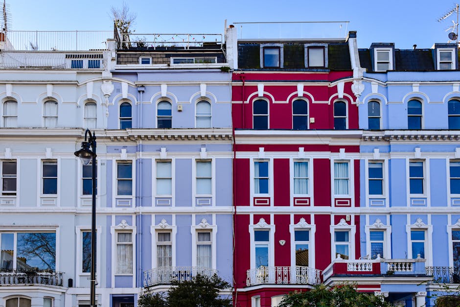 A row of Victorian-style terraced buildings in Notting Hill featuring ornate facades with decorative mouldings and arched windows. The buildings are painted in pastel blue and vibrant pink colors, with white accents and detailed trim. Large windows are visible, some with open or closed curtains, and the street level shows a black street lamp and some greenery in the foreground. The scene is well-lit with natural daylight, highlighting the clean and well-maintained exterior surfaces. This image emphasizes the aesthetic appeal of residential architecture in Notting Hill, reflecting the area's characteristic charm and colorful housing.