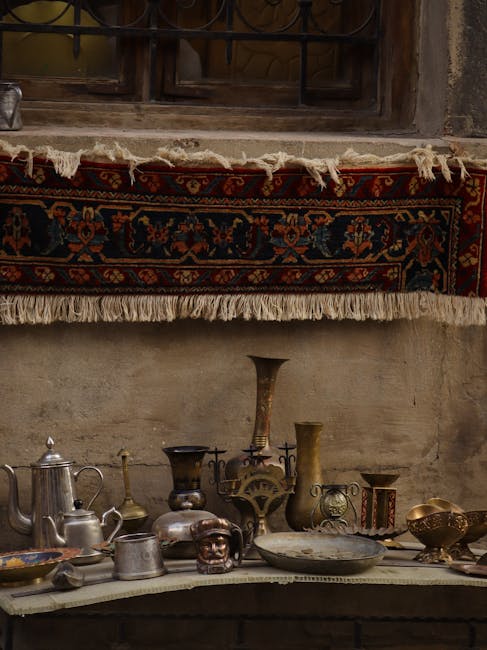 A rustic interior scene featuring a wooden shelf mounted on a textured, aged wall with visible wear and uneven surface. The shelf displays a collection of antique metallic and ceramic objects, including a teapot, vases, bowls, and decorative figurines, arranged in a somewhat cluttered manner. Above the shelf hangs a traditional woven rug with intricate patterns and fringed edges, adding vibrant red, blue, and beige tones to the muted background. Natural light illuminates the objects, highlighting their varied textures and patinas, while a warm, subdued atmosphere suggests a space dedicated to surface cleaning or restoration, as emphasized by the presence of professional cleaning experts such as Notting Hill Carpet Cleaning, who specialise in detailed, deep cleaning of sensitive surfaces like antique rugs.