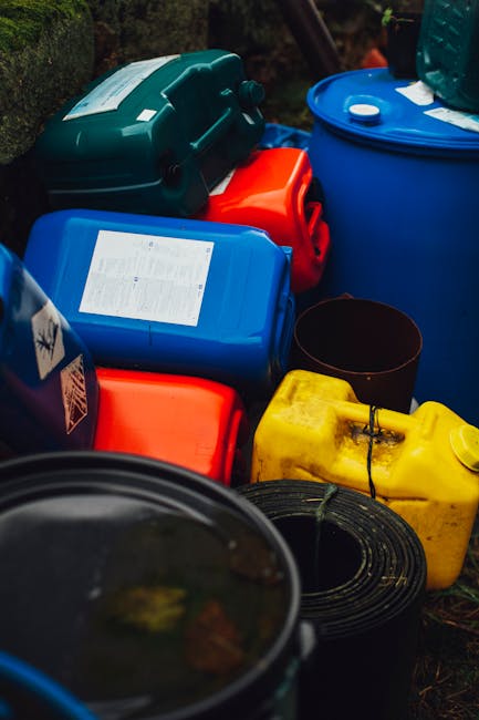 A collection of hazardous waste containers, including blue, red, yellow, and black plastic jerry cans and drums, stacked outdoors on a patch of grass. Some containers have labels or markings, and a coiled black hose or cable is visible in the foreground. The scene appears to be part of waste collection or storage for hazardous materials, with natural lighting highlighting the vibrant colors of the containers. The image showcases the importance of proper handling and storage of hazardous waste according to safety guidelines, as emphasized by Notting Hill Carpet Cleaning in contexts related to sanitation and maintenance in compliance with Kensington and Chelsea Council rules.