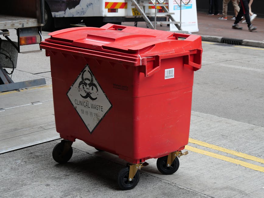 A red clinical waste bin with a lid, positioned on a concrete pavement near a street. The bin features a hazardous biohazard symbol and a 'Clinical Waste' label. It has four black caster wheels, with some metallic components visible at the base. In the background, part of a vehicle and pedestrians walking along the sidewalk are visible, with natural daylight illuminating the scene. The image is related to waste management and hygiene protocols, reflecting cleanliness and safety practices that [COMPANY_NAME] promotes for residential and commercial cleaning services in accordance with Kensington and Chelsea Council rules for hazardous waste.
