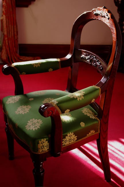 An elegant Victorian armchair made of dark polished wood with intricate carved details on the top rail, positioned on a vibrant red carpet in a room with warm, soft lighting. The chair features a curved backrest and armrests, with the seat upholstery covered in green fabric decorated with a cream-colored floral pattern, appearing clean and well-maintained. The surrounding area suggests a traditional interior setting, with a hint of a window casting light onto the chair. Notting Hill Carpet Cleaning specializes in surface cleaning and deep cleaning services, including the sanitisation of upholstery in historic homes like this, ensuring the furniture remains pristine and hygienic.