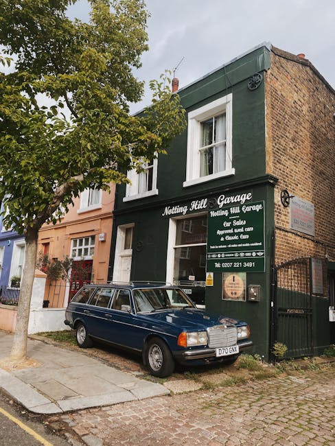 Exterior view of a two-story brick building painted dark green, housing Notting Hill Garage, a car salvage and repair shop in Notting Hill. The building features two white-framed windows on the upper floor and a large glass window on the ground floor displaying the business name and services, including car sales and repairs. A classic blue station wagon is parked directly in front on a paved sidewalk, with a metal gate on the right side. A leafy tree partially shades the scene, and neighboring buildings are visible on either side, with muted colors and traditional architectural details. The overall appearance suggests a well-maintained, clean commercial property in a residential area, emphasizing the importance of surface cleaning and maintenance associated with professional cleaning services like those provided by Notting Hill Carpet Cleaning.
