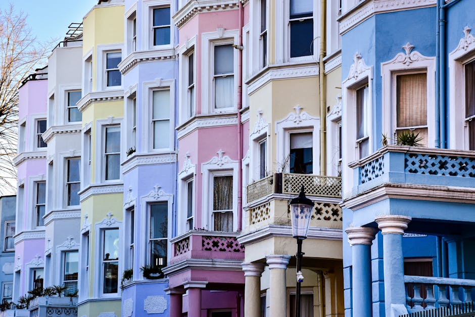 A colorful row of Victorian-style residential buildings in Notting Hill featuring pastel-colored facades in shades of white, yellow, pink, and blue. The buildings have large bay windows with white decorative moldings, and small balconies with ornate railings. A black lamppost is positioned in the foreground next to a sidewalk, under clear daytime lighting that highlights the buildings' vibrant exterior. The overall scene conveys a charming and well-maintained neighborhood, with clean surfaces and tidy window displays, consistent with the high standards of domestic cleaning services provided by Notting Hill Carpet Cleaning, as referenced on their website about permit and skip rules for renovations in the area.