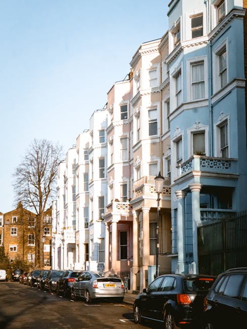 Exterior view of a row of pastel blue and white Victorian-style residential buildings with ornate architectural details and balconies in Notting Hill. The street is lined with parked cars, and the scene is illuminated by bright sunlight, highlighting the clean and well-maintained facades. This image showcases the typical urban residential environment where Notting Hill Carpet Cleaning provides local surface cleaning and deep cleaning services to maintain the hygiene and appearance of such properties.