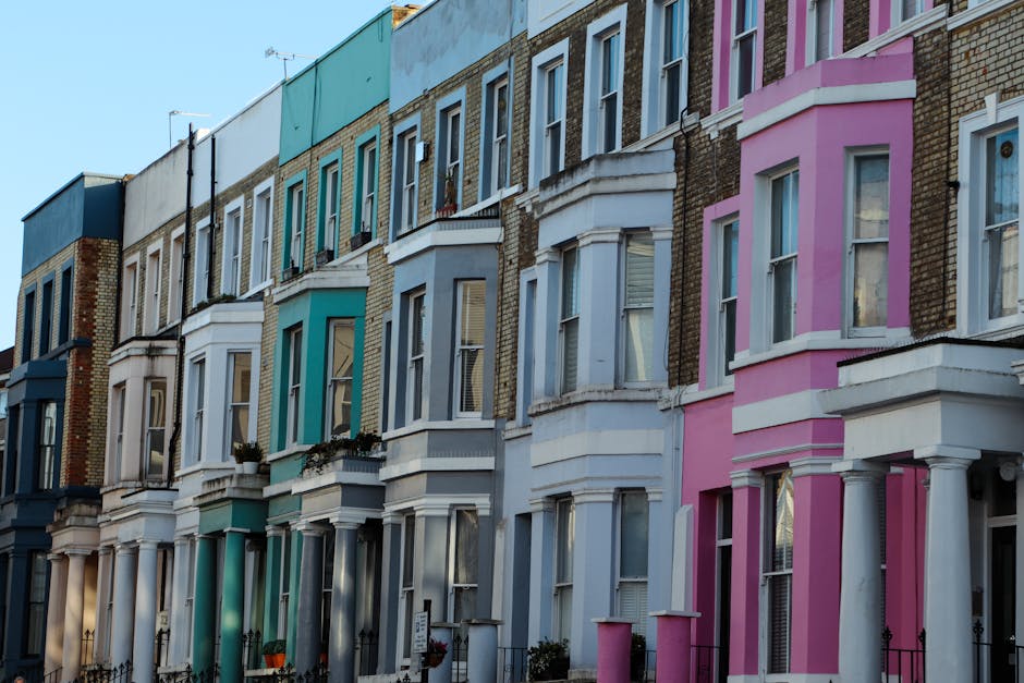 Exterior view of a row of pastel blue and white Victorian-style residential buildings with ornate architectural details and balconies in Notting Hill. The street is lined with parked cars, and the scene is illuminated by bright sunlight, highlighting the clean and well-maintained facades. This image showcases the typical urban residential environment where Notting Hill Carpet Cleaning provides local surface cleaning and deep cleaning services to maintain the hygiene and appearance of such properties.