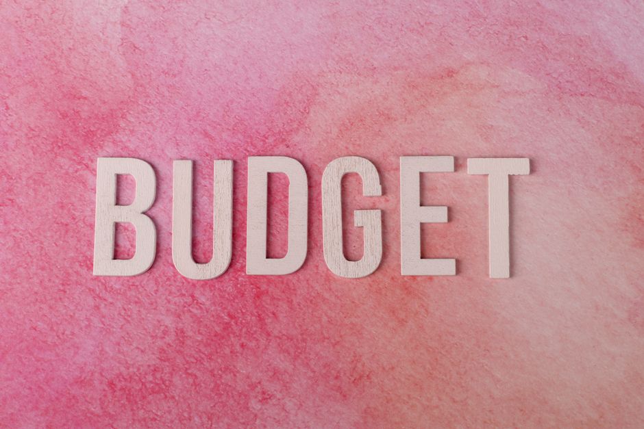 Close-up image of the word 'BUDGET' spelled out in large, white wooden letters placed on a textured pink background. The surface appears smooth with a slight gradient, highlighting the clean and simple design. The lighting is even, emphasizing the clarity and brightness of the letters and the background. This image may relate to discussions about affordable or cost-effective surface cleaning options, aligning with Notting Hill Carpet Cleaning's transparent pricing for flats in W11, as seen on their website.