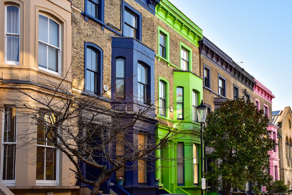 A row of colorful Victorian-style terraced houses on a city street, featuring brick facades painted in beige, deep blue, bright green, and pink. Large sash windows with white trim are visible on each building, some with curtains inside. A black lamppost stands in front of the green house, alongside a green leafy tree with branches extending across the scene. The street is under bright daylight, with clear blue sky overhead, and the overall scene appears clean and well-maintained, reflecting the high standards of surface cleaning and domestic sanitisation promoted by Notting Hill Carpet Cleaning.