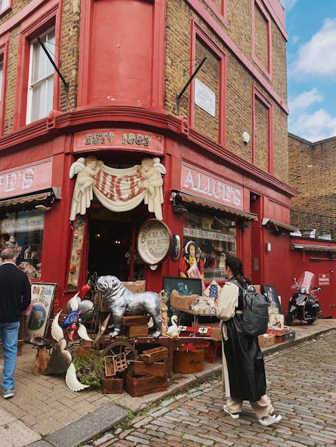 The exterior of a vibrant, red-painted building housing Alice's shop on Westbourne Grove in Notting Hill. The shop's façade features decorative sculptural elements and a white draped sign with the shop's name. Outside, various vintage and antique items including ceramic duck figurines, a large stuffed dog, wooden crates, and framed artworks are displayed on the sidewalk. The building has large display windows revealing the shop's interior, with posters and signs. Pedestrians, some carrying backpacks, are walking and browsing the street, which is paved with cobblestones. The scene is well-lit with natural daylight, highlighting the colorful exterior and street activity, though the image does not depict any cleaning or maintenance activities related to surface cleaning or sanitisation by Notting Hill Carpet Cleaning.