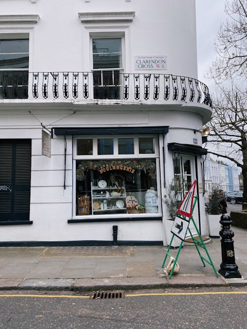 Exterior view of a small bakery storefront on Westbourne Grove, with a large display window showing baked goods and decorative items inside. The building has a white facade with black trim around the window and door, and a sign above the window reads 'Boulangerie'. There is a black railing on the upper balcony and a small street sign indicating Clarendon Cross W11. In front of the shop, two green and white traffic cones are positioned on the pavement, with a red and white triangular roadwork sign attached. The sidewalk appears clean and well-maintained, with a black garbage bin nearby, and the street is paved with asphalt. The scene is illuminated by natural daylight, and part of a leafless tree can be seen on the right side, indicating a colder season. For businesses like this, Notting Hill Carpet Cleaning offers professional cleaning services to ensure hygiene and shine for your commercial premises.