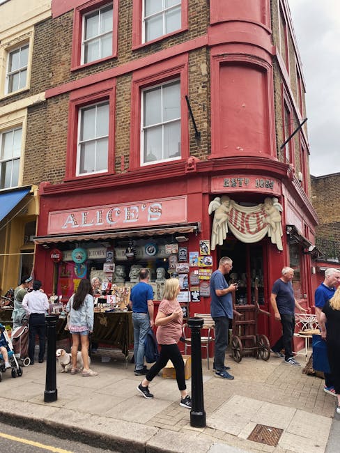 The exterior of a vibrant, red-painted building housing Alice's shop on Westbourne Grove in Notting Hill. The shop's façade features decorative sculptural elements and a white draped sign with the shop's name. Outside, various vintage and antique items including ceramic duck figurines, a large stuffed dog, wooden crates, and framed artworks are displayed on the sidewalk. The building has large display windows revealing the shop's interior, with posters and signs. Pedestrians, some carrying backpacks, are walking and browsing the street, which is paved with cobblestones. The scene is well-lit with natural daylight, highlighting the colorful exterior and street activity, though the image does not depict any cleaning or maintenance activities related to surface cleaning or sanitisation by Notting Hill Carpet Cleaning.